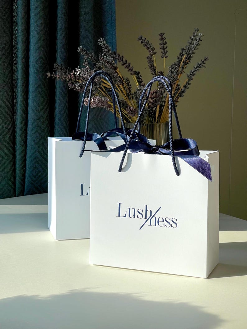 May include: Two white gift bags with the word "Lushness" printed in navy blue. The bags have navy blue handles and are tied with navy blue ribbons. Dried lavender is in a clear vase in the background.