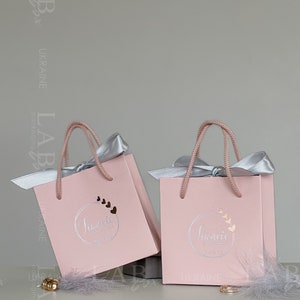 May include: Two pink gift bags with silver handles and bows. The bags have a logo that says "Jumelle" with a circle of hearts around it. The bags are on a white surface with a gray feather.