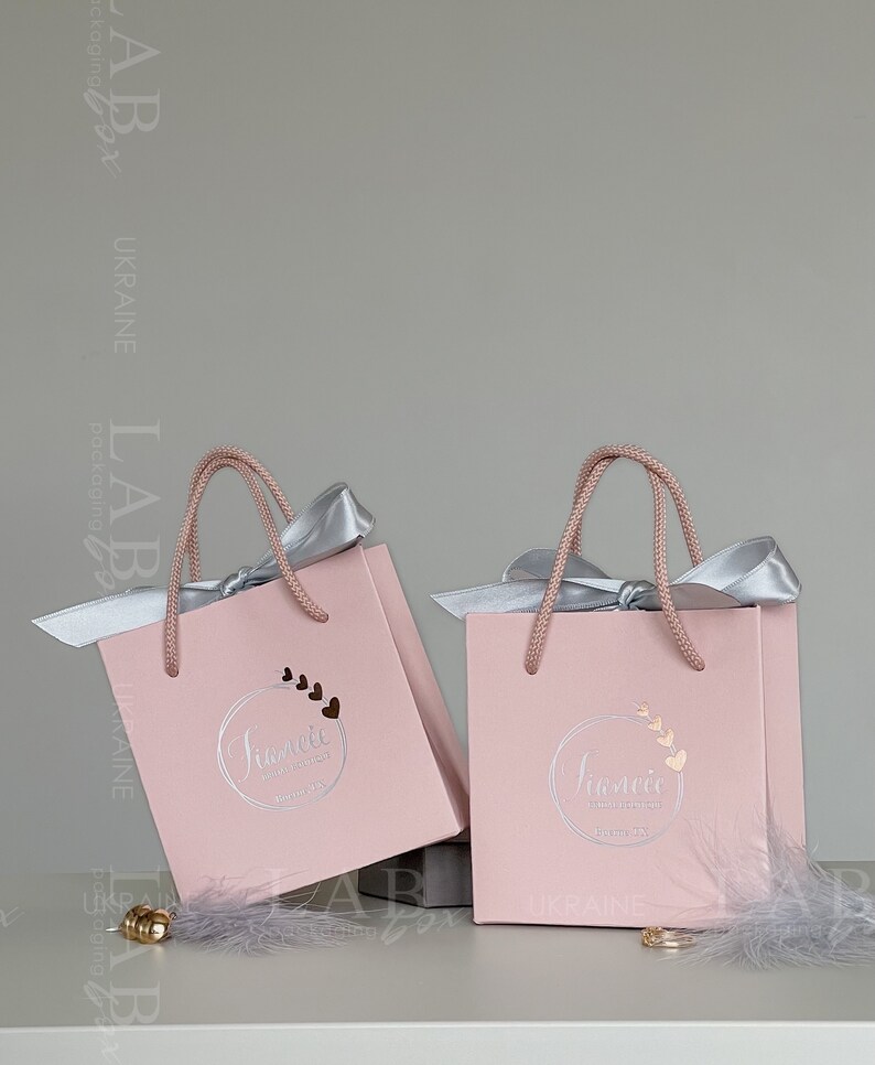 May include: Two pink gift bags with silver handles and bows. The bags have a logo that says "Jumelle" with a circle of hearts around it. The bags are on a white surface with a gray feather.