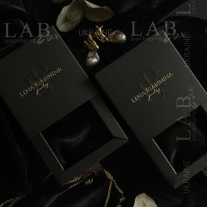 May include: Two black jewelry boxes with gold lettering that reads "LAB packaging box" and "LENA RIABININA jewelry". The boxes are on a black velvet surface with a gold chain necklace and a sprig of eucalyptus.