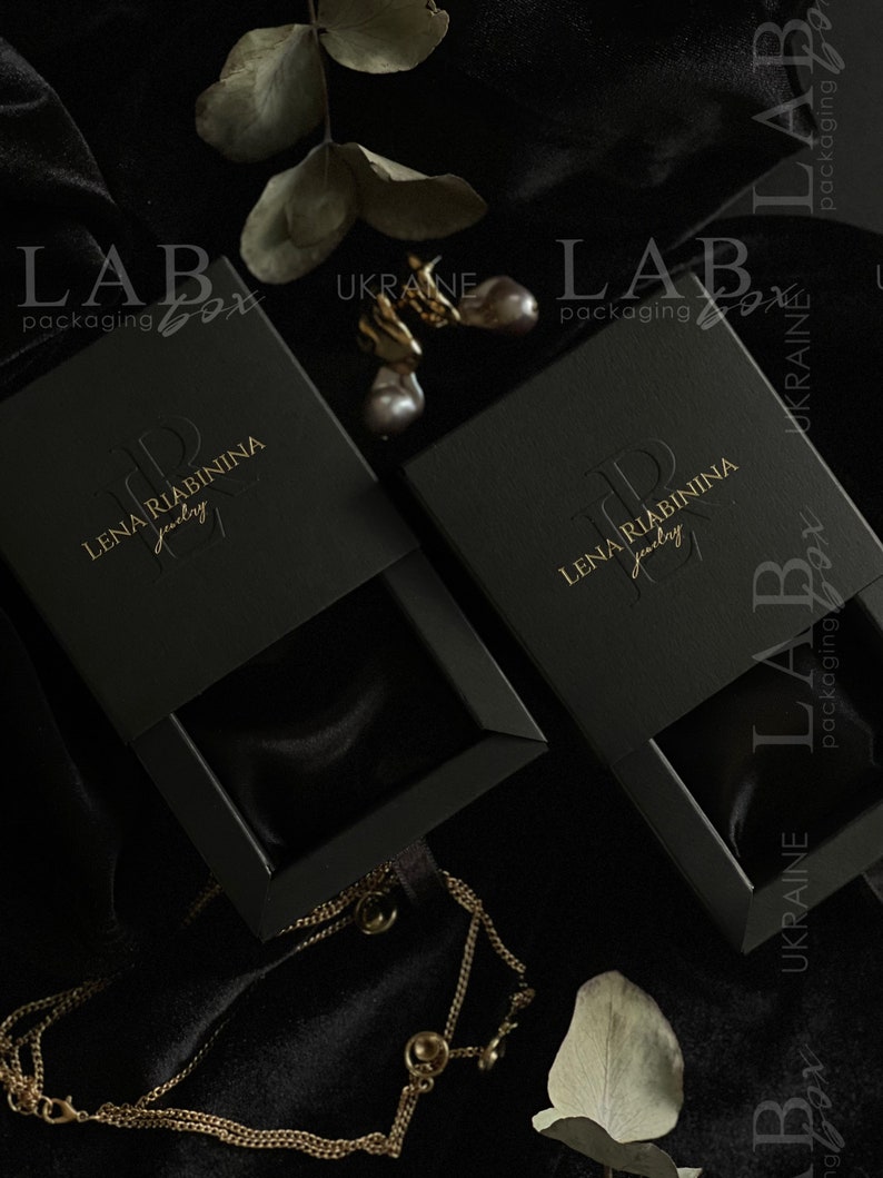 May include: Two black jewelry boxes with gold lettering that reads "LAB packaging box" and "LENA RIABININA jewelry". The boxes are on a black velvet surface with a gold chain necklace and a sprig of eucalyptus.