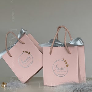 May include: Two pink gift bags with silver handles and silver ribbons. The bags have a white circle logo with the text "Fianc&eacute;e Bridal Boutique" and the city "Boerne, TX".