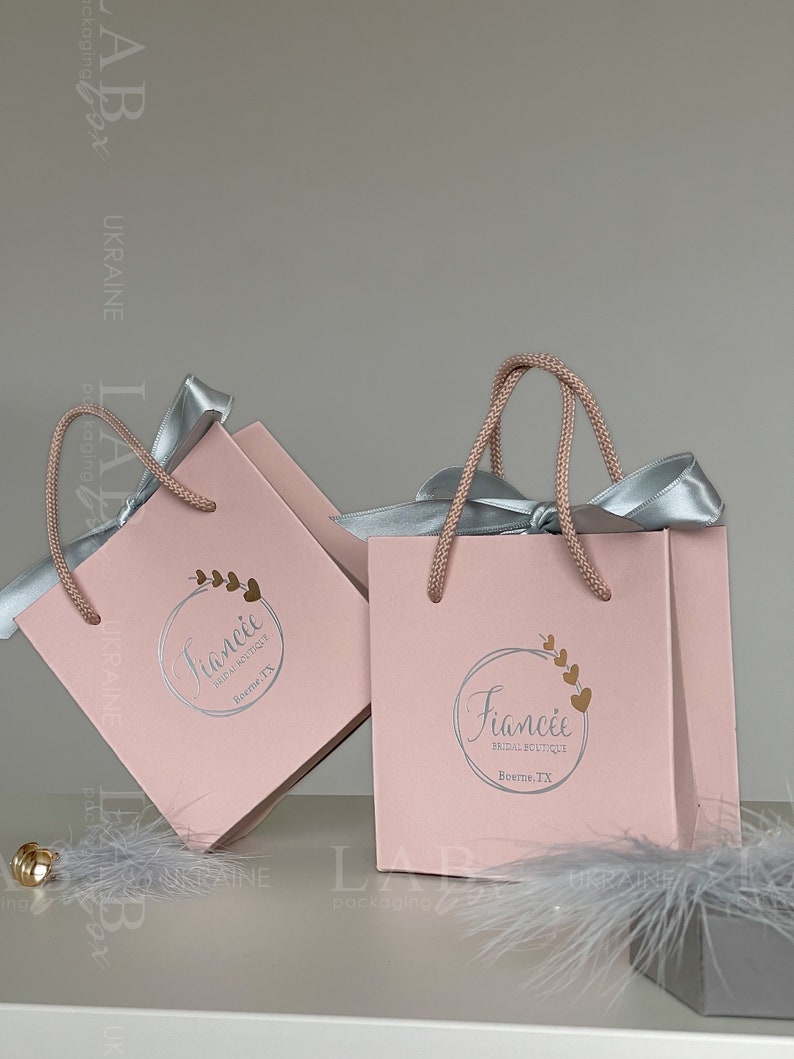 May include: Two pink gift bags with silver handles and silver ribbons. The bags have a white circle logo with the text "Fianc&eacute;e Bridal Boutique" and the city "Boerne, TX".