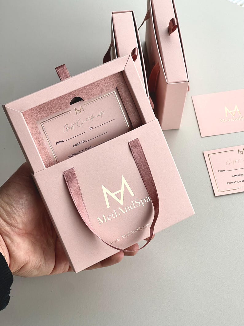May include: Pink gift certificate box with a matching gift card. The box has a ribbon handle and contains a gift certificate with the text "Gift Certificate". The logo "M MedAndSpa" is visible on the box and card.