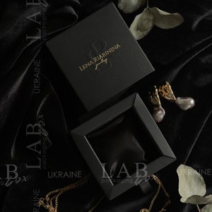 May include: A black jewelry box with a gold logo that reads "Lena Riabinin Jewelry". The box is open and a gold chain necklace is lying on a black velvet surface.