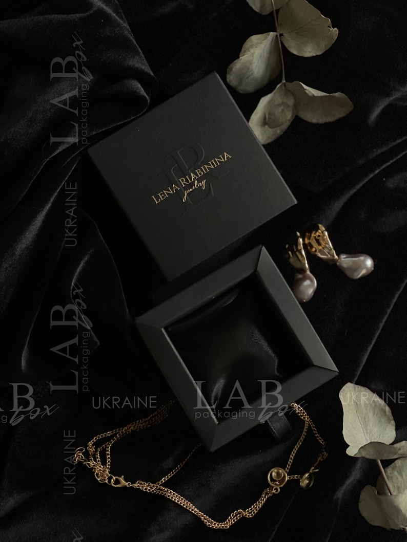 May include: A black jewelry box with a gold logo that reads "Lena Riabinin Jewelry". The box is open and a gold chain necklace is lying on a black velvet surface.