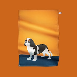May include: A kitchen towel featuring a black and white illustration of a Cavalier King Charles Spaniel dog on a yellow background with a blue border.