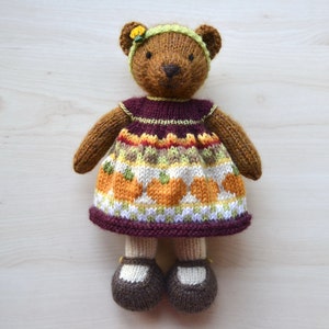 May include: A brown knitted teddy bear wearing a burgundy dress with a pumpkin pattern. The dress has white, orange, and green accents. The bear is wearing brown shoes.