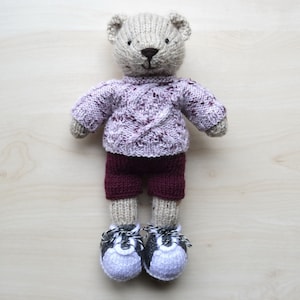 May include: A knitted teddy bear wearing a purple sweater with a white cable knit pattern and maroon pants. The bear is wearing white and black sneakers.