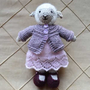 May include: A knitted toy lamb wearing a purple cardigan and a pink lace dress with purple shoes.
