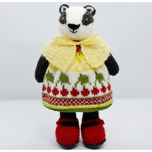 May include: A knitted badger doll wearing a yellow dress with a white and green pattern and red cherries. The doll is wearing a yellow cape with a yellow ribbon and red shoes.