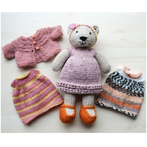 May include: A knitted teddy bear wearing a pink dress with a pink cardigan and a striped dress. The bear also has a pink flower headband and orange shoes.