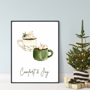 May include: Watercolor print featuring two mugs of hot chocolate. One mug is white with a whipped cream topping and the other is green with marshmallows. The words "Comfort & Joy" are written in cursive below the mugs. The print is in a black frame.