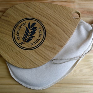 May include: A wooden cutting board with a teardrop shape and a handle. The board has a black logo that says "Magic Wood Valburden" with a stylized leaf design. The board is sitting on a white cloth bag.