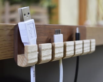 May include: A wooden cable organizer with six slots for holding cords and wires. The organizer is mounted on a wooden surface and has a natural wood finish. There are three white cords and one black cord in the slots.