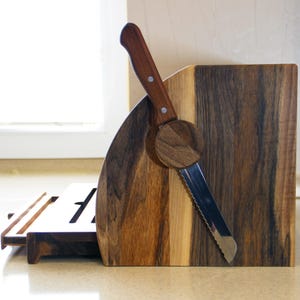 Puede incluir: Una caja de pan de madera con un cuchillo para pan incorporado. La caja est&aacute; hecha de madera oscura con un borde de madera clara. El cuchillo est&aacute; hecho de acero inoxidable y tiene un mango de madera.