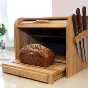 Op de afbeelding: Houten broodtrommel met een roldeksel, een uitschuifbare snijplank en een messenhouder. Een brood met sesamzaadjes ligt erin. Messen met houten handvatten zitten in de houder. De broodtrommel is van lichtgekleurd hout.