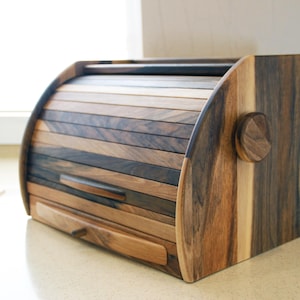 Puede incluir: Una caja de pan de madera con una tapa con bisagras y un caj&oacute;n. La caja est&aacute; hecha de diferentes tipos de madera, creando un patr&oacute;n &uacute;nico. La tapa est&aacute; hecha de listones de madera que se deslizan para abrirse y cerrarse.