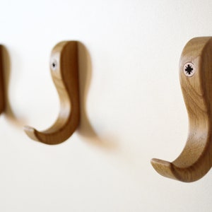 May include: Three wooden hooks mounted on a white wall. The hooks are shaped like a curved 'S' and have a natural wood finish.