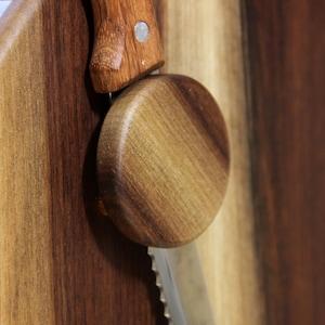 Puede incluir: Una caja de pan de madera con un soporte redondo para cuchillo de pan de madera fijado en el lateral. El soporte para cuchillo de pan est&aacute; hecho de madera oscura y tiene un patr&oacute;n de veta de madera clara.