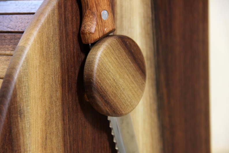 Puede incluir: Una caja de pan de madera con un soporte redondo para cuchillo de pan de madera fijado en el lateral. El soporte para cuchillo de pan est&aacute; hecho de madera oscura y tiene un patr&oacute;n de veta de madera clara.