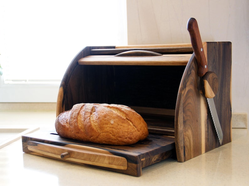 Puede incluir: Una caja de pan de madera con una tabla de cortar extra&iacute;ble y un soporte para cuchillos. La caja de pan est&aacute; hecha de madera oscura con un borde de madera clara. Una hogaza de pan est&aacute; sobre la tabla de cortar.