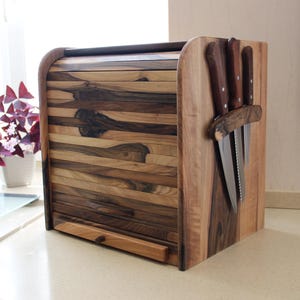 May include: A rectangular wooden bread box with a roll-top door featuring horizontal slats of varying brown wood tones. The right side of the unit includes a built-in magnetic rack holding three stainless steel kitchen knives with brown handles.