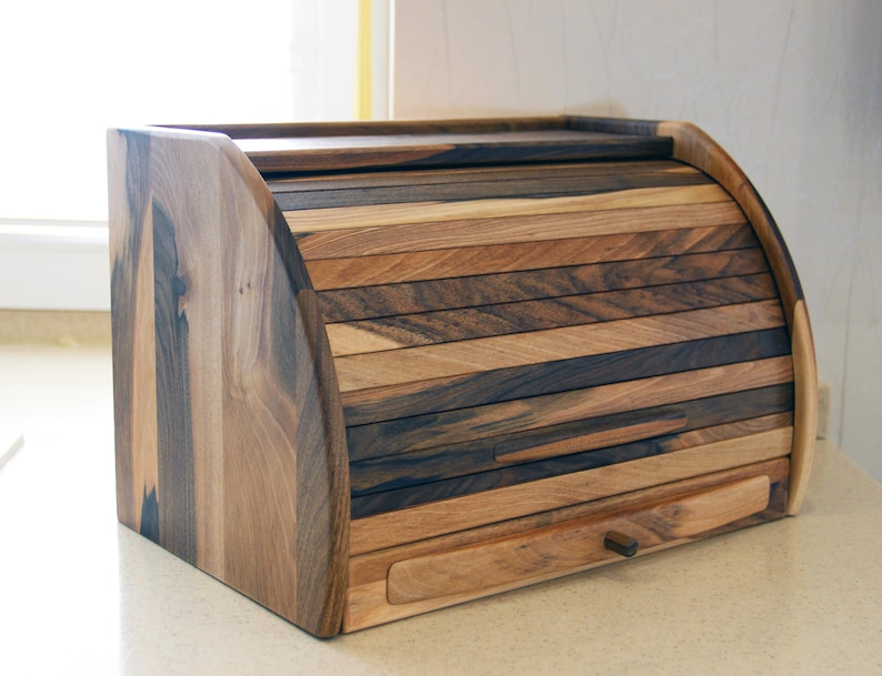 Puede incluir: Una caja de pan de madera con una tapa con bisagras y un caj&oacute;n. La caja est&aacute; hecha de madera oscura con un acabado natural. La tapa est&aacute; hecha de m&uacute;ltiples listones de madera que est&aacute;n dispuestos en un patr&oacute;n horizontal. El caj&oacute;n tiene una peque&ntilde;a manija de metal.