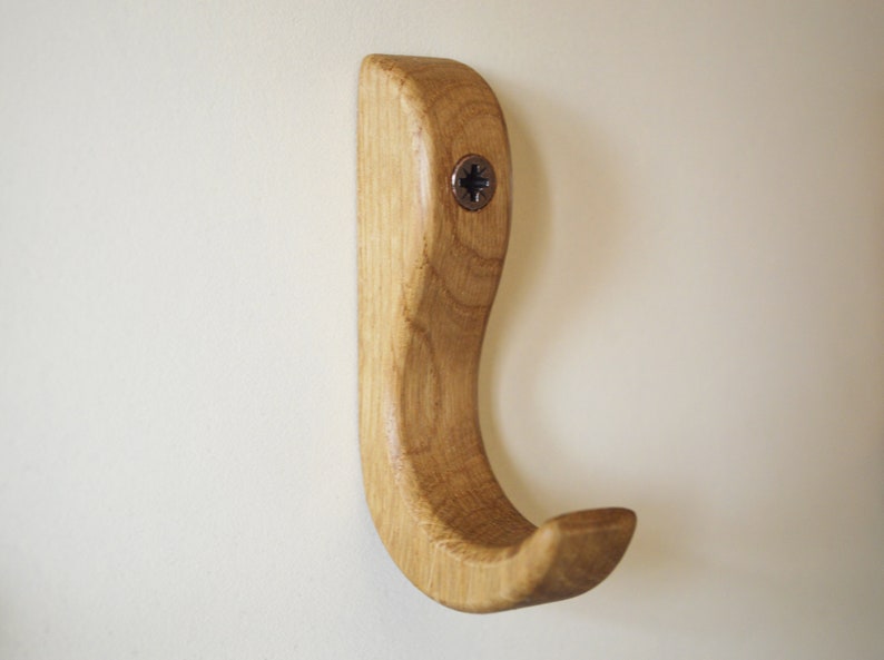 May include: A wooden hook with a curved design, finished in a natural wood tone. The hook is mounted to a wall with a screw.
