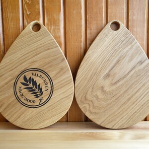 May include: Two wooden cutting boards in the shape of a teardrop with a hole in the top. The board on the left has a black circle with the text "MAGIC WOOD" and "VALBURDEN" around the edge. The board on the right is plain.