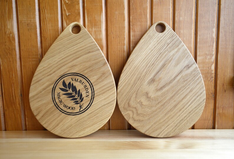 May include: Two wooden cutting boards in the shape of a teardrop with a hole in the top. The board on the left has a black circle with the text "MAGIC WOOD" and "VALBURDEN" around the edge. The board on the right is plain.