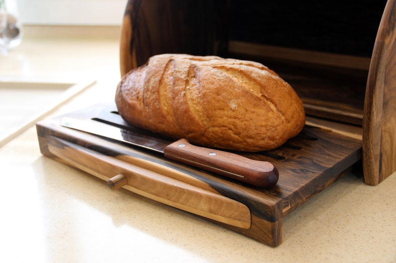 Puede incluir: Una caja de pan de madera con una hogaza de pan dentro y un cuchillo para pan en la tapa. La caja de pan est&aacute; hecha de madera oscura con un borde de madera clara.