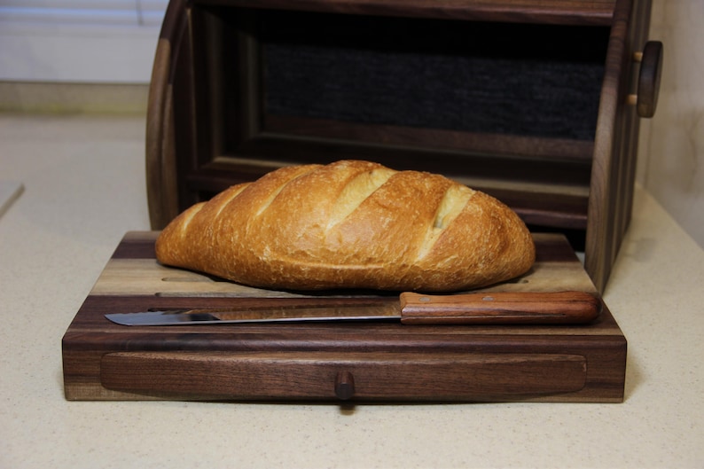 Puede incluir: Una tabla de cortar de madera con una hogaza de pan y un cuchillo con mango de madera. La tabla de cortar tiene un caj&oacute;n.