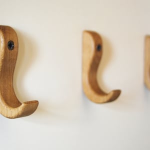 May include: Three wooden hooks mounted on a white wall. The hooks are a light brown color and have a simple, curved design.