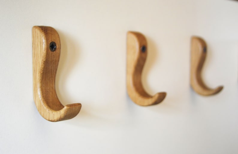 May include: Three wooden hooks mounted on a white wall. The hooks are a light brown color and have a simple, curved design.