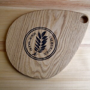 May include: A wooden cutting board with a teardrop shape and a handle. The board is made of light wood with a dark wood grain pattern. The board has a logo burned into the surface that says "Magic Wood Valburden".