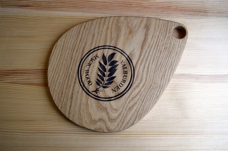 May include: A wooden cutting board with a teardrop shape and a handle. The board is made of light wood with a dark wood grain pattern. The board has a logo burned into the surface that says "Magic Wood Valburden".