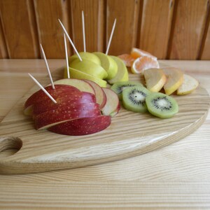 May include: A wooden serving board with a heart shape and a handle. The board is filled with sliced fruit, including red apples, green apples, kiwi, and oranges.