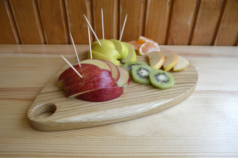 May include: A wooden serving board with a heart shape and a handle. The board is filled with sliced fruit, including red apples, green apples, kiwi, and oranges.