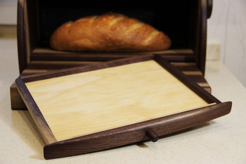 Puede incluir: Una caja de pan de madera con una bandeja extra&iacute;ble. La caja de pan est&aacute; hecha de madera oscura con detalles de madera clara. La bandeja est&aacute; hecha de madera clara con un marco de madera oscura. La caja de pan est&aacute; abierta y se puede ver una hogaza de pan en el interior.