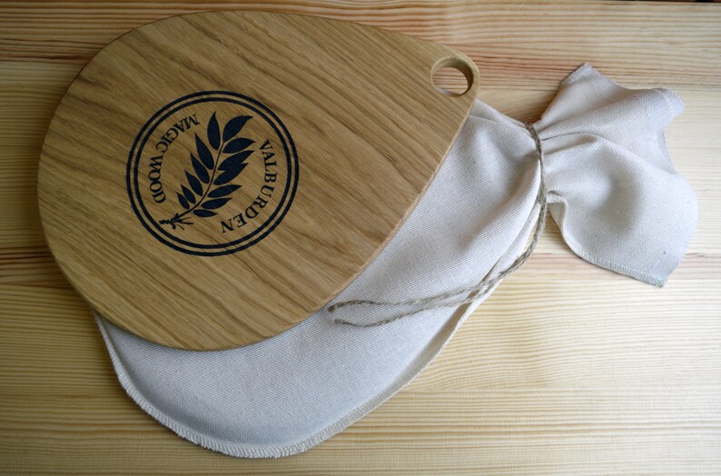 May include: A wooden cutting board with a teardrop shape and a handle. The board has a black logo that says "Magic Wood Valburden" with a stylized leaf design. The board is sitting on a white cloth bag.