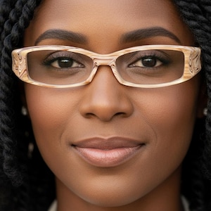 Op de afbeelding: Een close-up portret van een persoon met een lichtbruine rechthoekige bril met decoratieve details. De persoon heeft een donkere huid en zwart gevlochten haar. De achtergrond is een vage neutrale kleur.