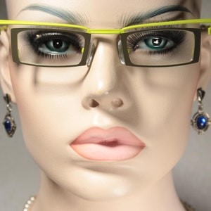 May include: Eyeglasses with a lime green upper frame and rectangular lenses. The glasses are on a mannequin with blue-green eyes and full lips. Dangling earrings with blue stones are visible.