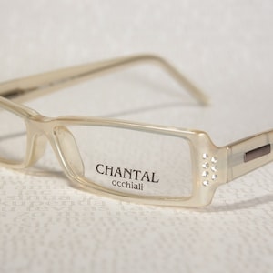 Peut inclure: Une paire de lunettes rectangulaires transparentes avec un accent brun sur le côté. Les lunettes ont des strass sur le côté et disent "CHANTAL occhiali" sur la lentille droite.