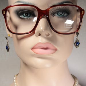 May include: A mannequin head wearing large, square, brown-framed eyeglasses with clear lenses. The glasses have a gold accent on the temples. The mannequin also wears blue gemstone earrings and a pearl necklace. The glasses have the word "ATELIER" on the lens.