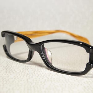 May include: Black eyeglasses with a rectangular frame and clear lenses. The temples are a light brown color. The glasses are set against a white background.