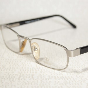 Könnte beinhalten: Silberne Brille mit schwarzen Bügeln und klaren Gläsern. Der Markenname "BRILKE" ist auf dem rechten Glas sichtbar.