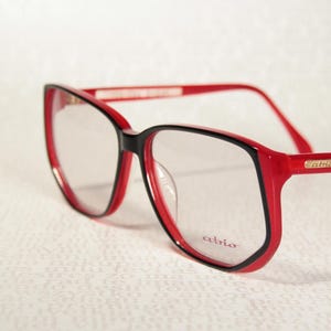 Op de afbeelding: Bril met een glanzend rood montuur en een zwarte binnenrand. De heldere glazen zijn voorzien van het woord atrio in het rood aan de rechterkant. Een klein goudkleurig rechthoekig plaatje met de merknaam atrio is zichtbaar op de brilpoot.