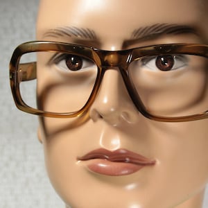 May include: A close-up of a mannequin wearing brown plastic framed glasses with a rectangular shape. The glasses have a brown tint and are worn on a mannequin with a neutral expression.