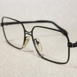 May include: A pair of spectacles with a thin black metal frame and a square-shaped lens design. The glasses feature clear lenses, adjustable nose pads with a light yellow tint, and slender black temples that extend from the outer corners.
